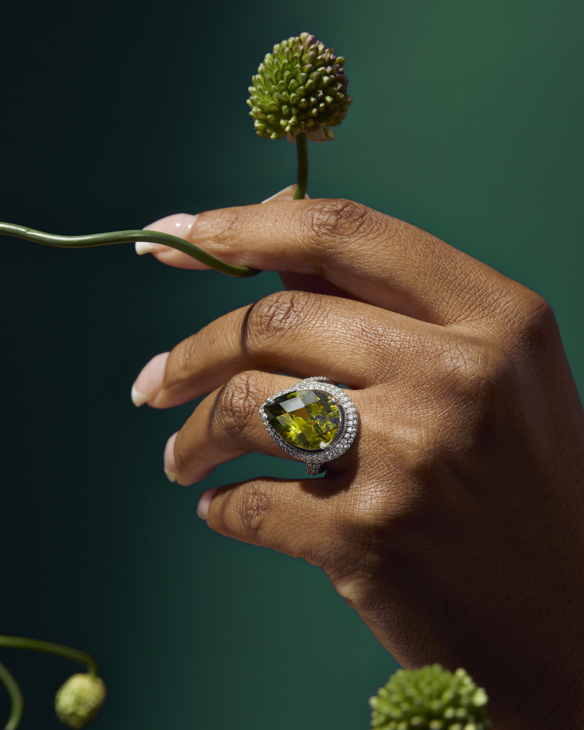 A close-up of a dark-skinned hand wearing a pear-cut olive green gemstone ring with a diamond halo, holding a small green flower.
