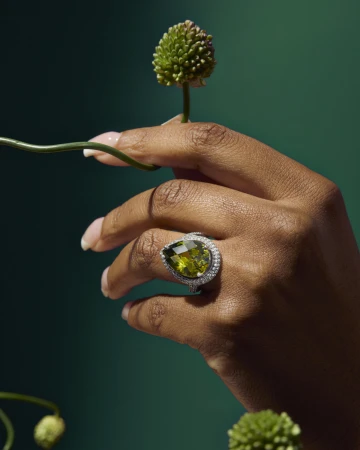 A close-up of a dark-skinned hand wearing a pear-cut olive green gemstone ring with a diamond halo, holding a small green flower.