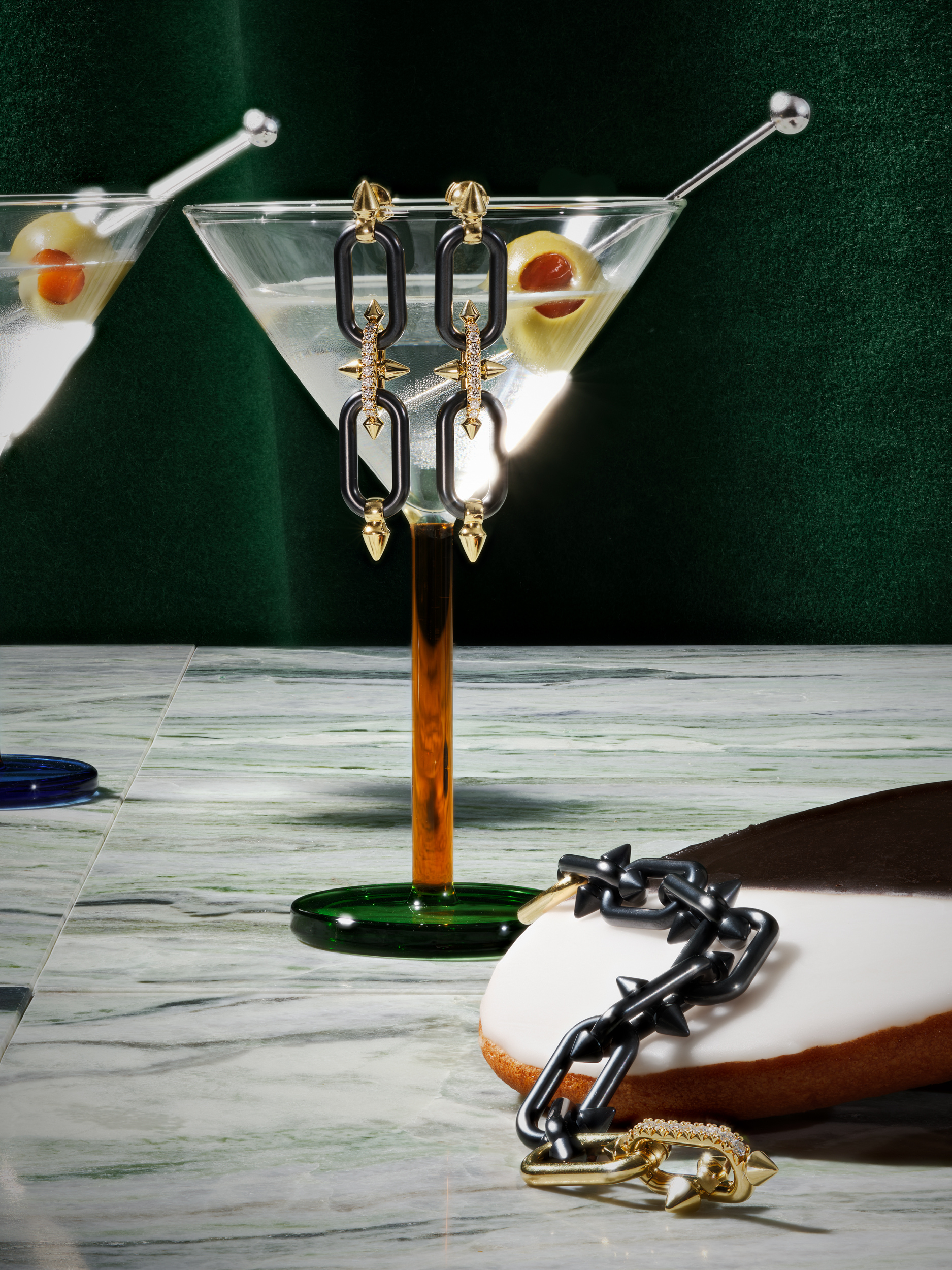 A pair of black chain link earrings suspended in front of a martini glass with green olives, next to a matching black chain link bracelet.