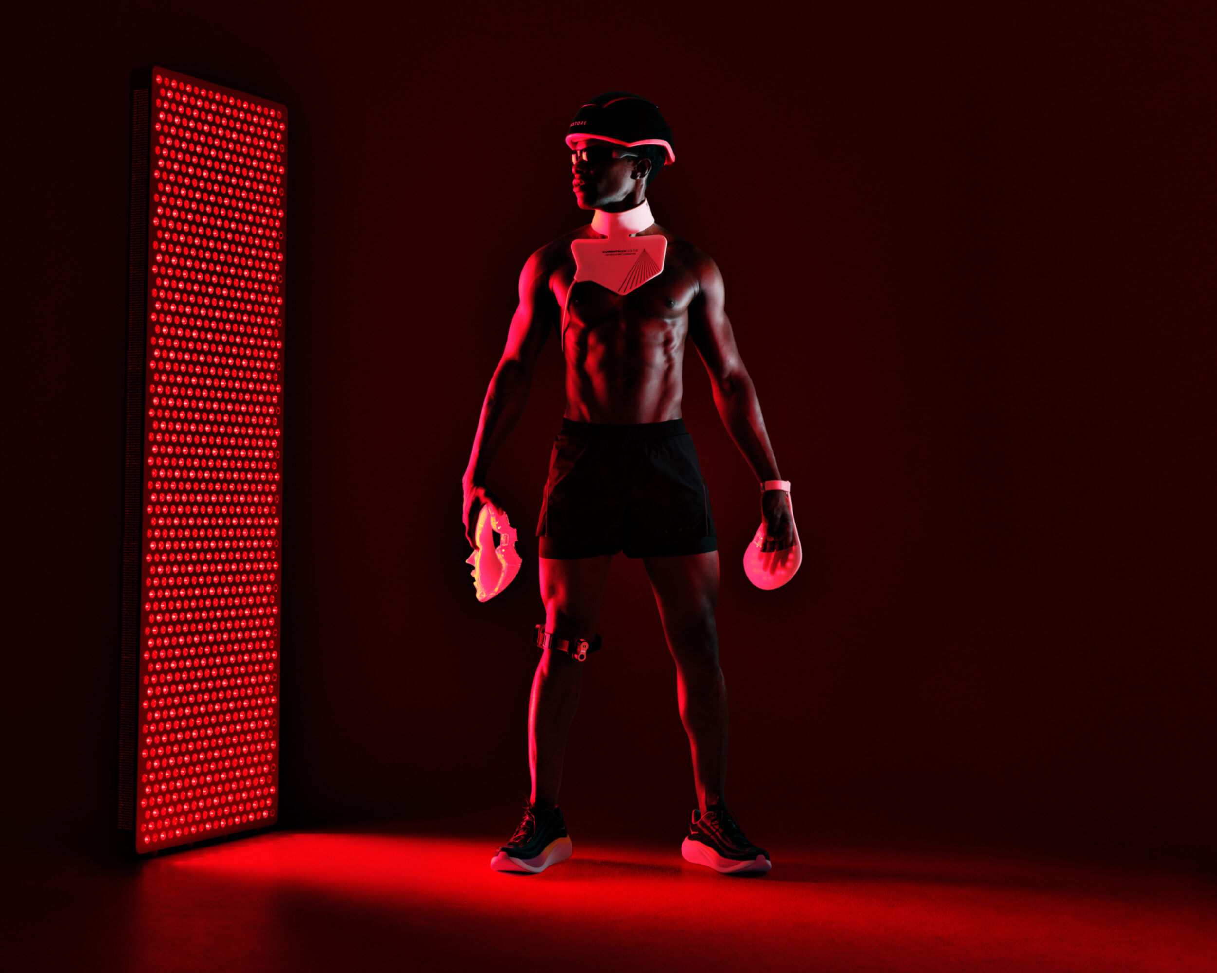 A Black man stands in profile wearing a futuristic helmet and chest plate, illuminated by a tall, glowing red LED therapy panel.