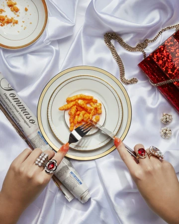 Close-up of hands with statement rings using a fork and knife to eat orange snack sticks off a fine china plate on white silk.