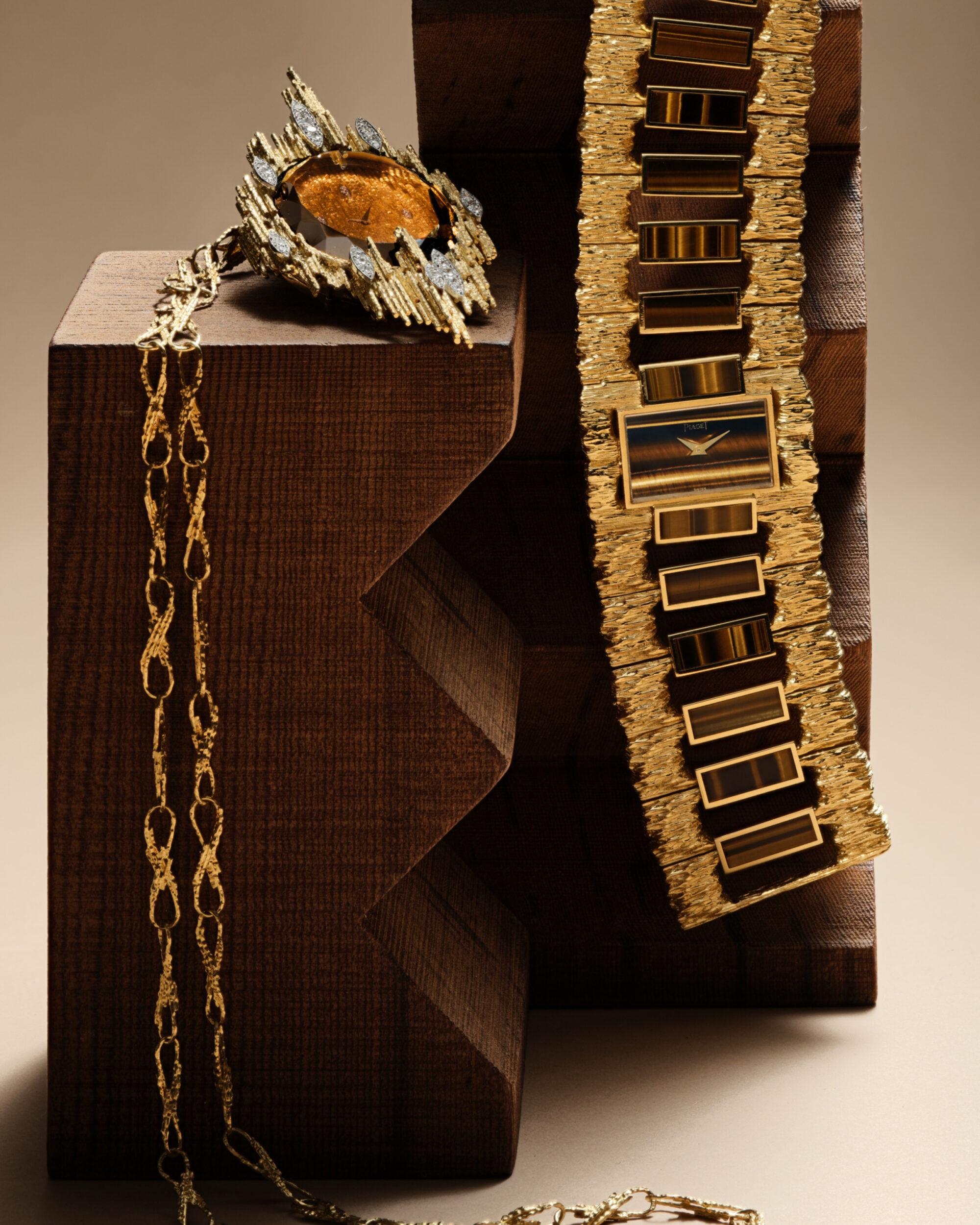 A gold watch with tiger eye inlays and a matching gold pendant necklace resting against a dark brown stepped wooden sculptural pedestal.