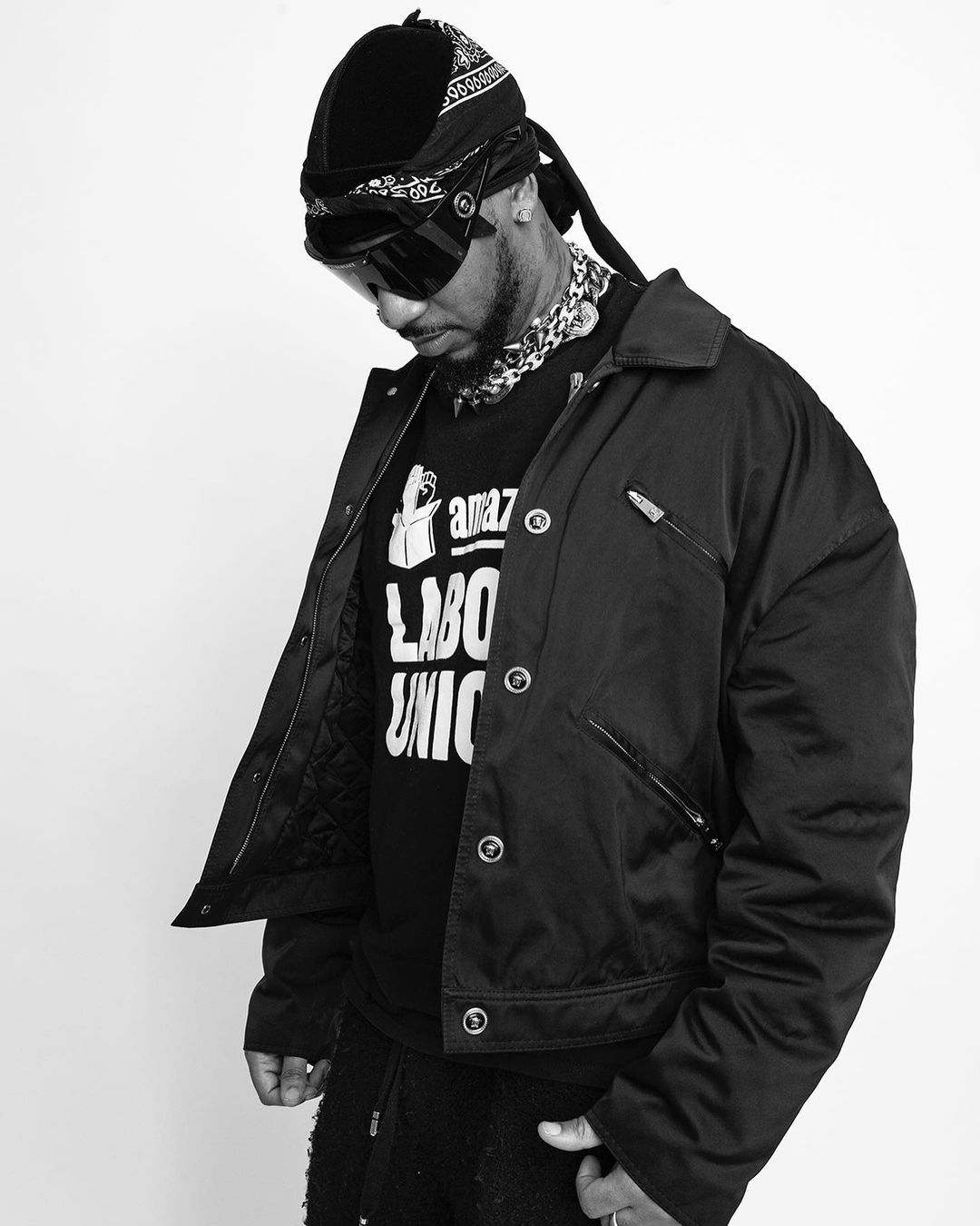 Black and white portrait of an adult male wearing a black jacket, t-shirt, head covering and sunglasses, looking down in a photo studio.