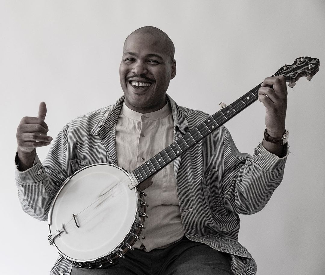 Black musician smiles while playing a banjo indoors. He is wearing a pinstripe jacket and giving a thumbs up to the camera.