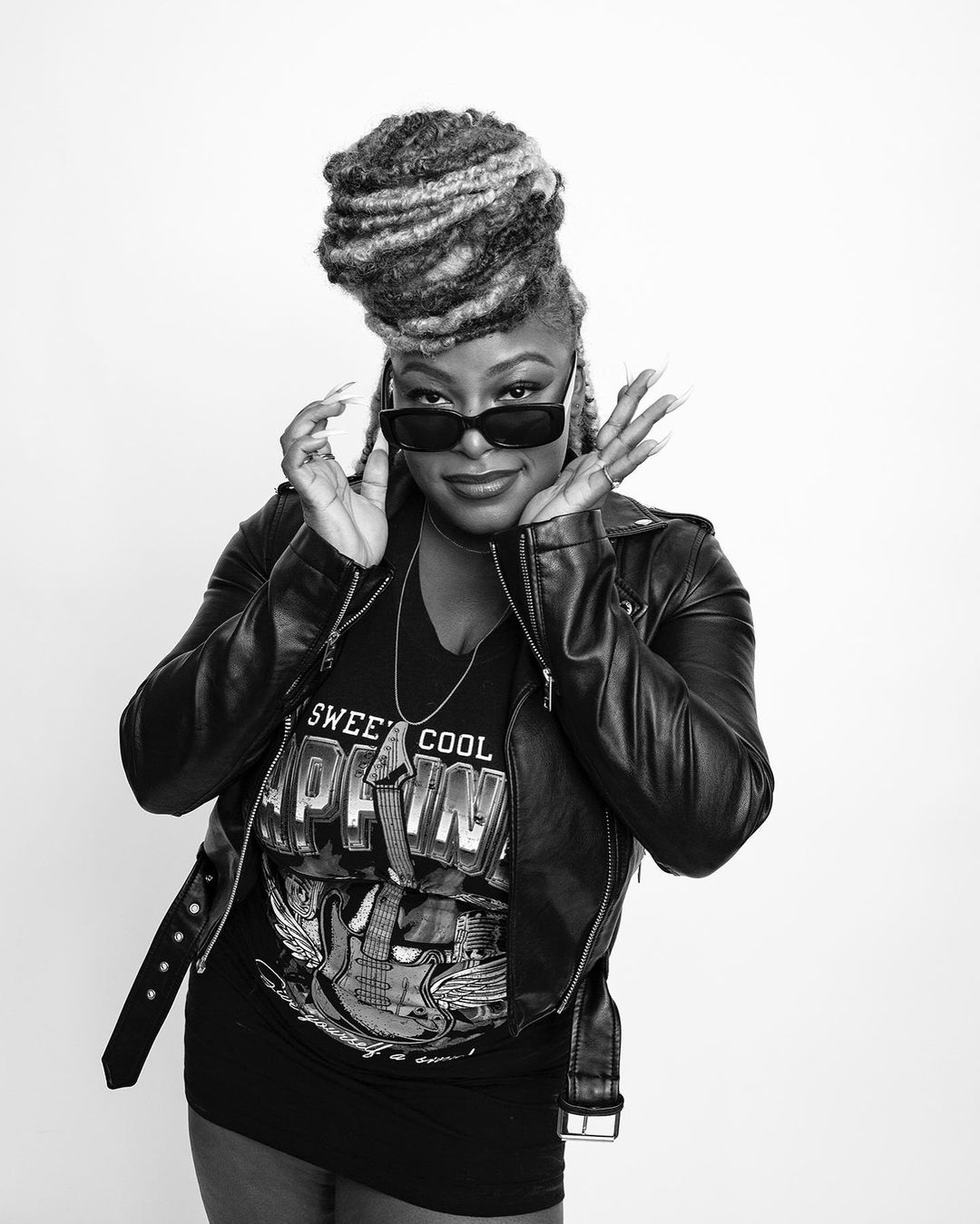 Black and white portrait of a stylish Black woman wearing sunglasses and a leather jacket, striking a pose against a neutral backdrop.