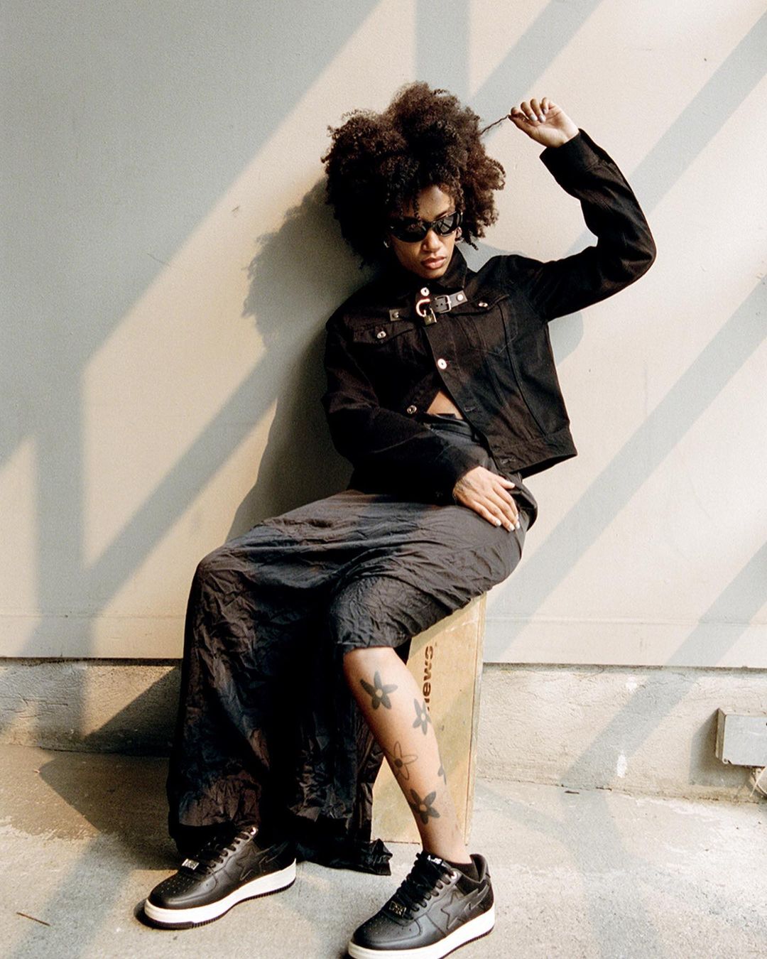 A woman with dark, curly hair poses in a black jacket and grey skirt, sitting on a box with black sneakers in an indoor setting.
