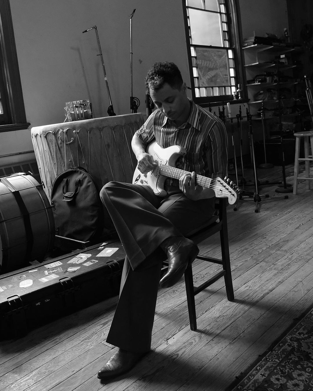 Black and white shot of a musician playing an electric guitar while seated on a chair in a music studio with vintage instruments.