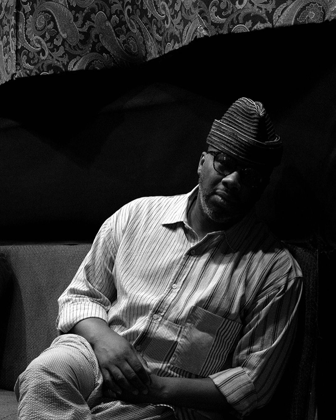 Monochrome portrait of a man in a striped shirt and hat, seated on a sofa with patterned drapery in the background; black and white.