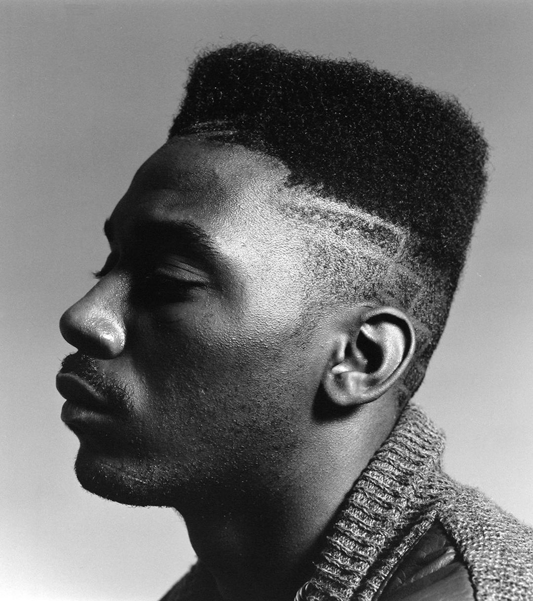 Black and white portrait of an adult Black man in profile wearing a cardigan, close up on the upper body, in a studio setting.