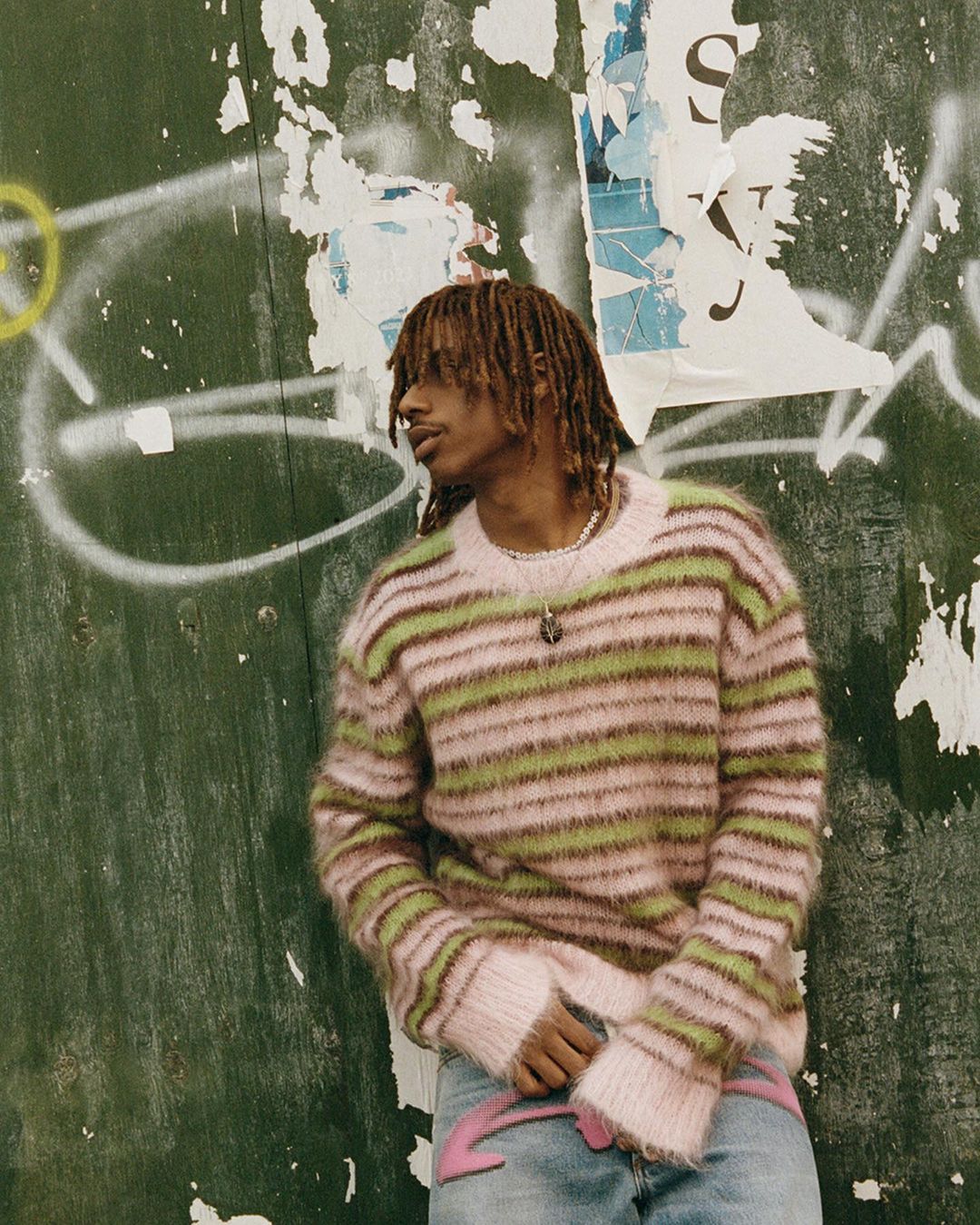 Portrait of a young Black man with locs wearing a pink and green striped sweater, leaning against a weathered wall with graffiti.