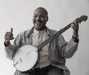 Black man smiling while holding a banjo and giving a thumbs up, photographed by Janette Beckman against a light background