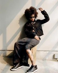 Portrait of a Black woman in a black jacket and long skirt sitting outdoors in London, photographed by Janette Beckman.