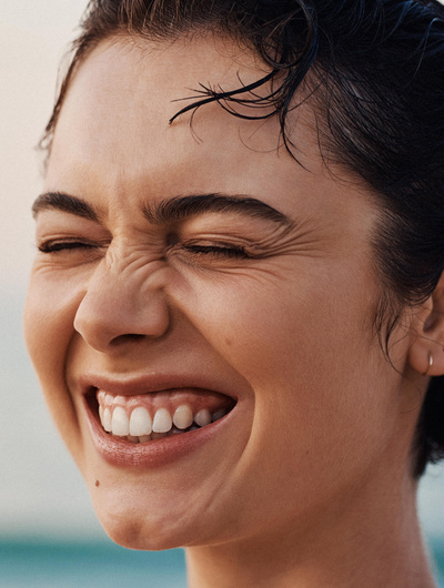 Close-up portrait of a fair-skinned woman with dark hair and a mole smiling with squinted eyes outdoors in bright, natural lighting.