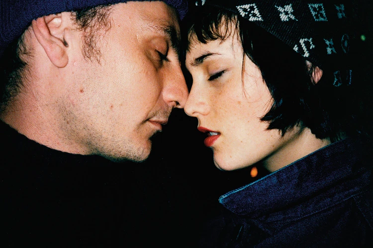 Close-up shot of a romantic couple. A fair-skinned man in a dark beanie with stubble touches noses with a fair-skinned woman.