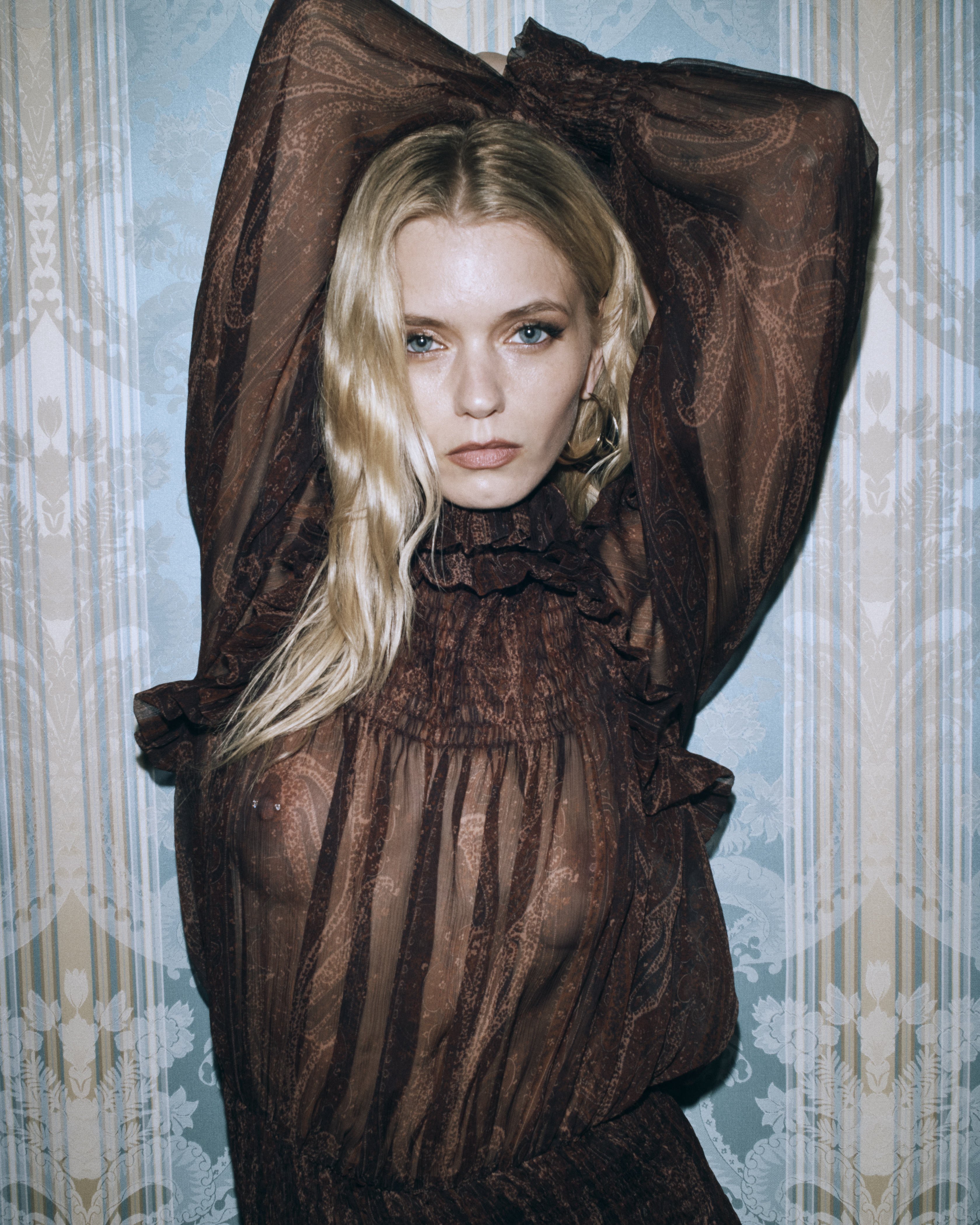 Portrait of a blonde woman with arms raised, wearing a brown sheer paisley blouse, posing in front of a blue paisley wallpaper.
