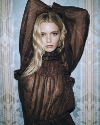 Blonde woman wearing a sheer brown patterned blouse with her arms raised against blue patterned wallpaper