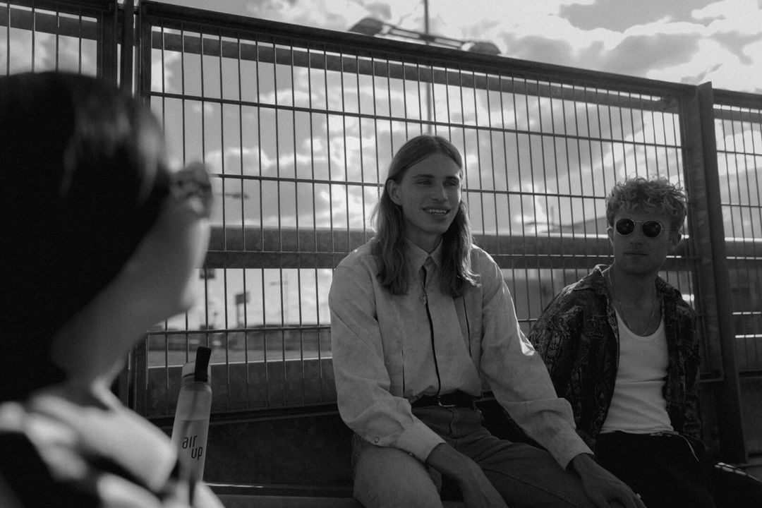 Black and white shot of three young adults sitting outdoors against a metal fence, with the sky and clouds in the background.