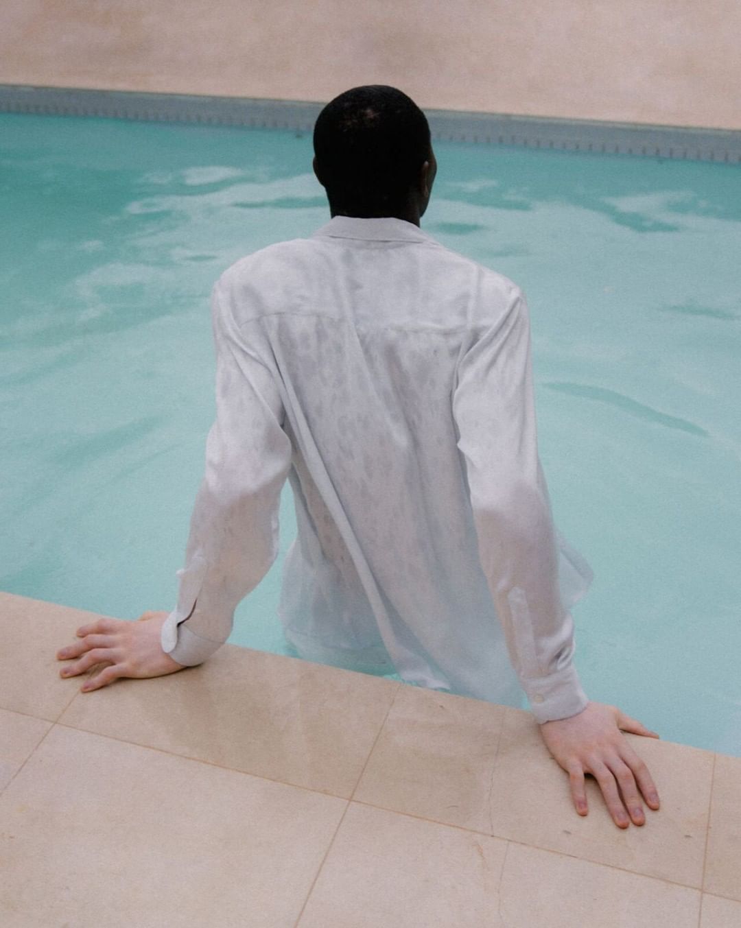 A person in a wet, white button-down shirt is standing in a swimming pool and facing away from the camera, with their hands on the pool's edge.