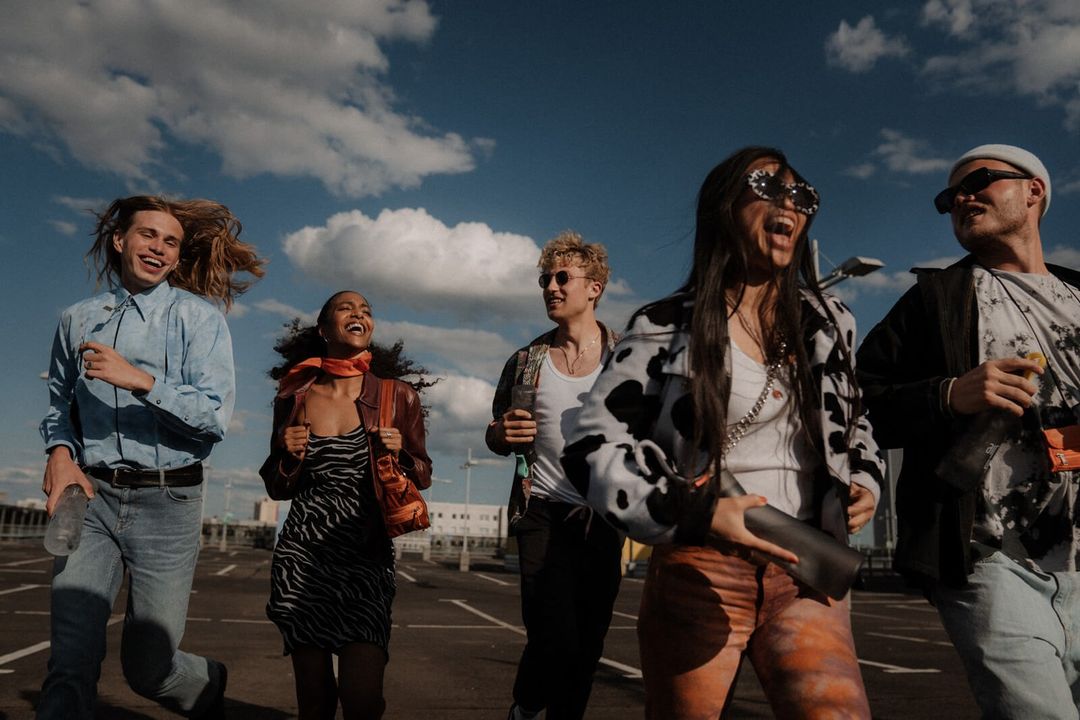 Group of five young adults smiling and walking on a rooftop parking lot under a partly cloudy sky, bathed in sunlight.