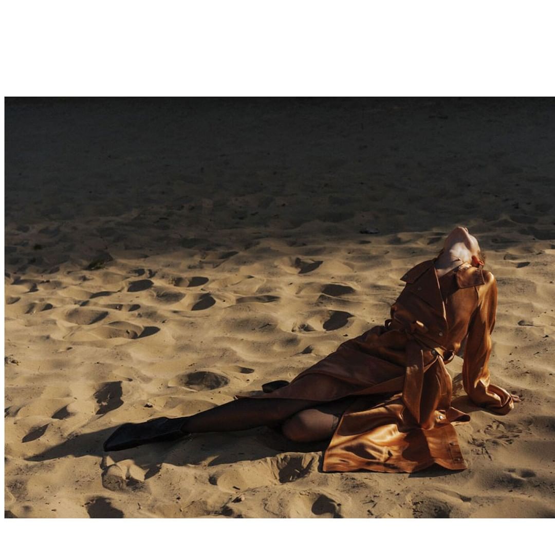 Woman in orange satin trench coat lies on a sandy beach, with one leg visible in black hosiery and dark boots, under soft light.