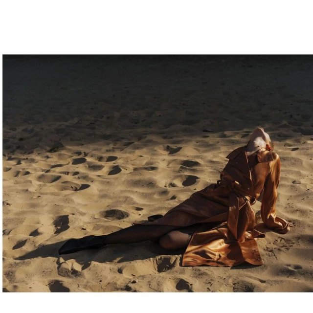 Woman in orange satin trench coat lies on a sandy beach, with one leg visible in black hosiery and dark boots, under soft light.