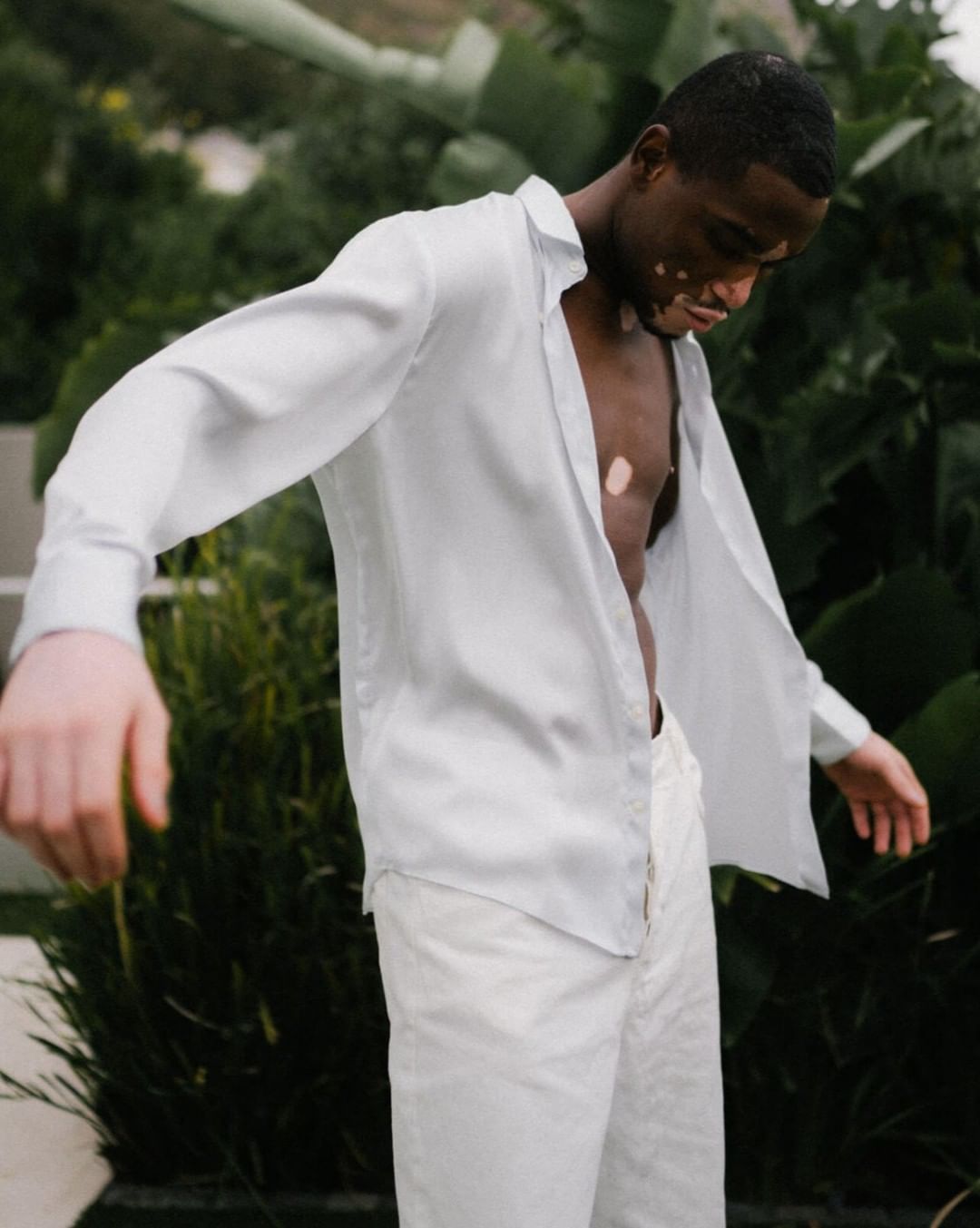 An African-American man wearing a white shirt and white pants poses outdoors in front of green tropical foliage, showcasing a fashion-forward look.
