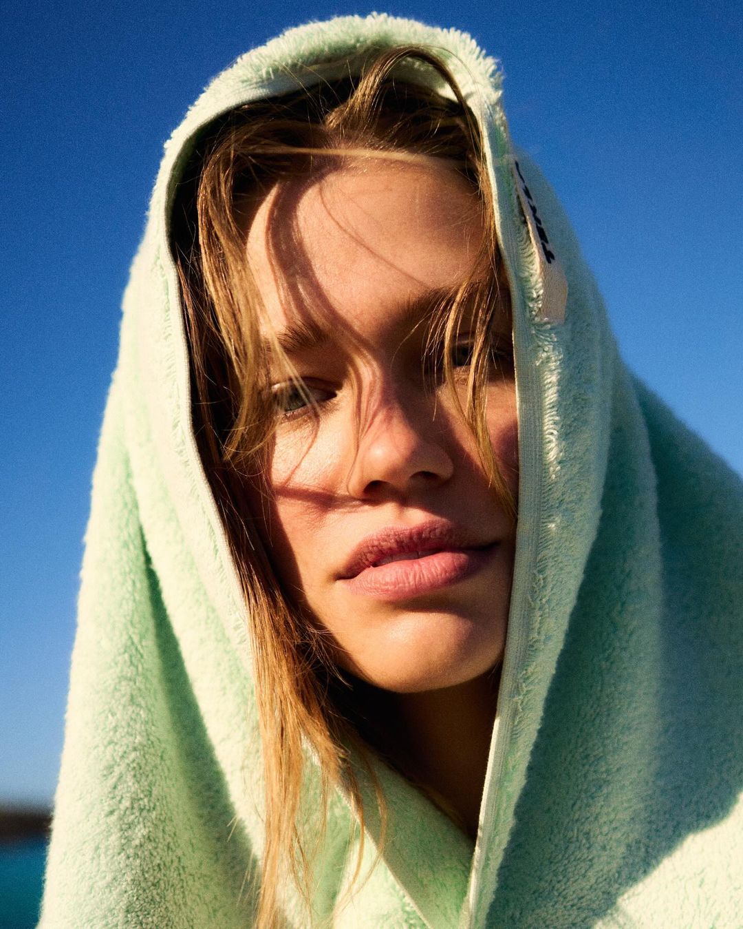 Close-up portrait of a young woman with blonde hair wrapped in a light green towel against a blue sky, bathed in sunlight.