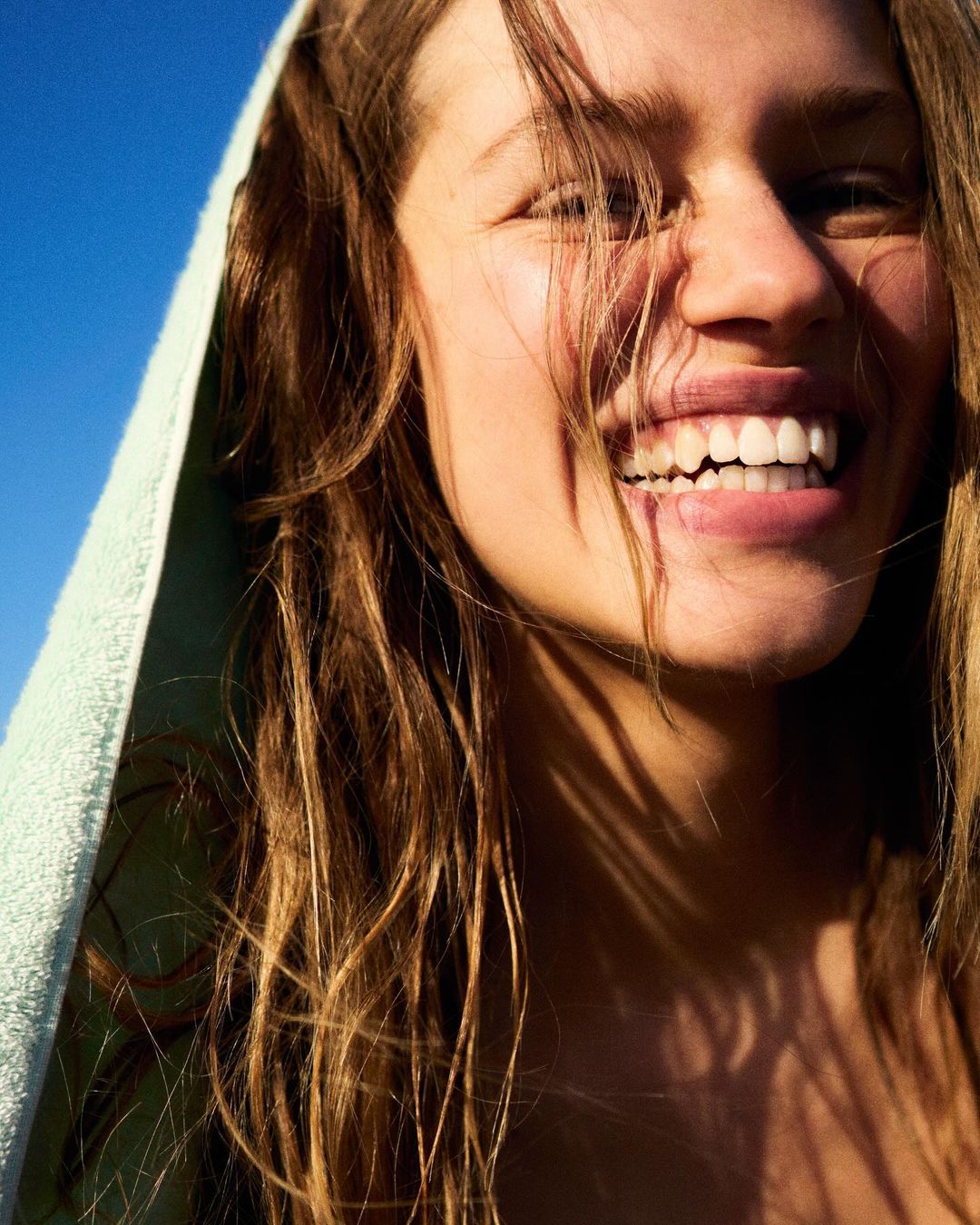 Close-up of a smiling woman with long brown hair and a towel draped over her head, set against a bright blue sky.