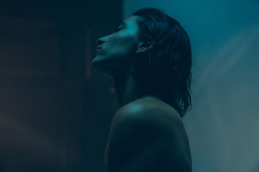 Profile of a shoulder-length dark-haired person illuminated by blue light, looking up in an indoor setting.