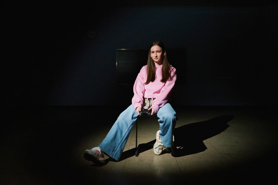 Portrait of a young woman with long brown hair sitting on a chair in jeans and a pink sweatshirt in a dark indoor space.