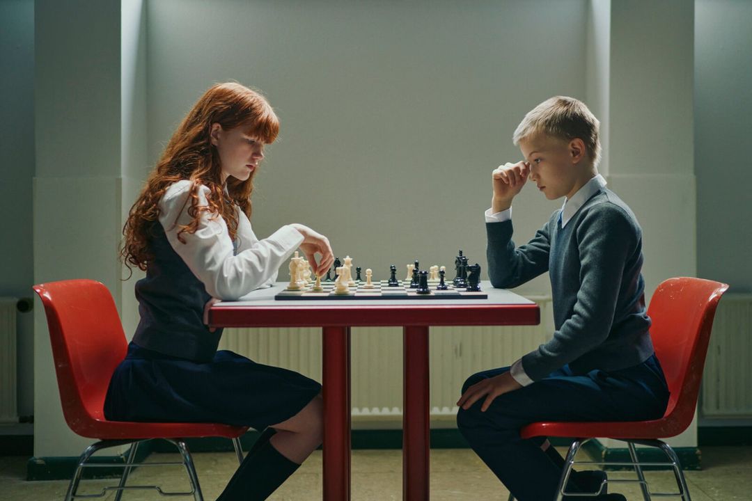 A red-haired girl and a blonde-haired boy play chess across a table in a bright room with red chairs, white walls, and recessed lighting.