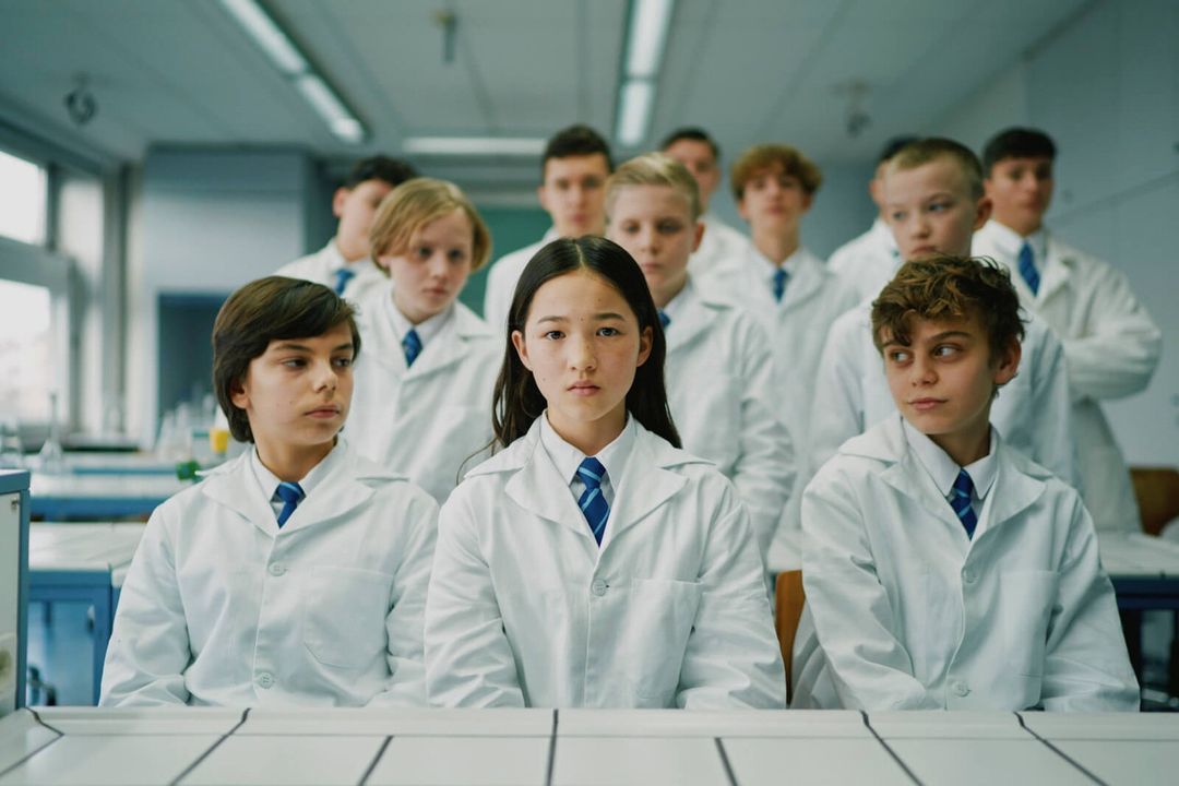 A diverse group of preteen children wearing matching white lab coats and ties sit at desks in a science classroom setting.