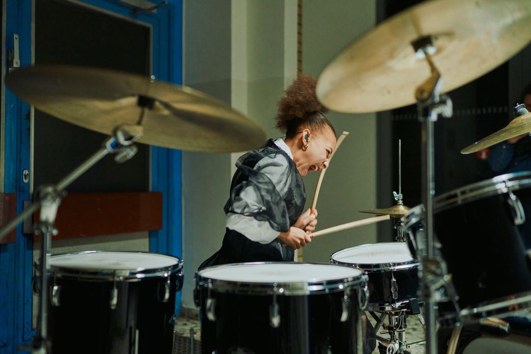 Energetic young female drummer enthusiastically playing a drum set indoors, showcasing her musical talent and passion in a vibrant performance.