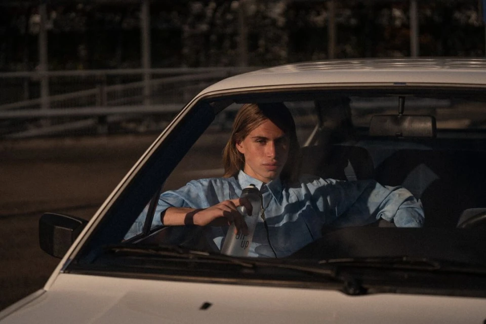 Man with long hair in a light-blue button-down shirt sits in a white car, holding a gray reusable bottle in an outdoor setting.