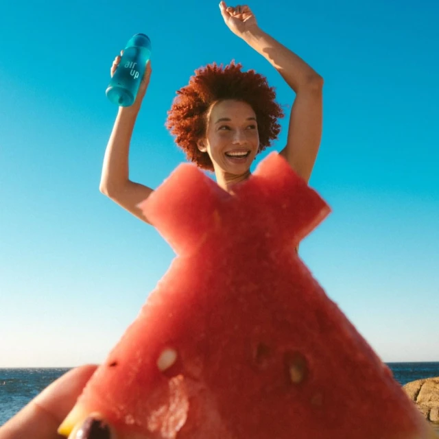 Happy woman holds a blue water bottle and a watermelon slice 'dress' against a bright blue sky, evoking summer fun and healthy hydration.