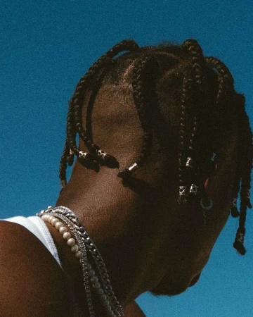 Stylish man with braids and pearl necklace against a serene blue sky. Fashionable jewelry and hairstyle in an outdoor setting.