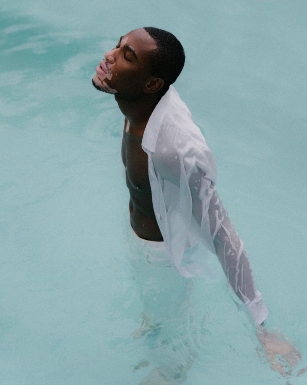 Young adult Black man in a white dress shirt stands chest-deep in pool water with his eyes closed, in a medium shot.
