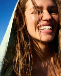 Close-up portrait of a smiling young woman with wet brown hair wrapped in a light green towel under a clear blue sky
