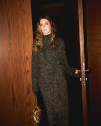 Woman in a dark sparkly knit dress holding a gold handbag in a doorway by Janine Sametzky