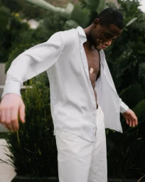 Black man in an unbuttoned white shirt and white trousers standing outdoors near green foliage, photographed by Janine Sametzky in Berlin.