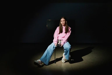 Young woman in a pink hoodie and light blue jeans seated on a chair in a dark room under a spotlight, photographed by Janine Sametzky in Berlin.