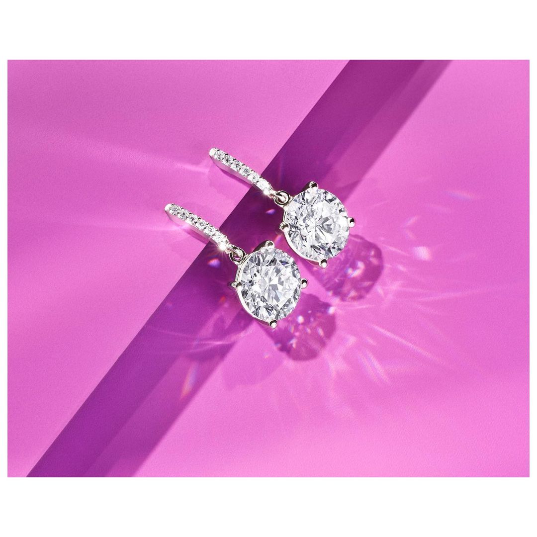 Two round-cut diamond drop earrings are displayed on a purple background with visible shadows, highlighting their brilliant sparkle.
