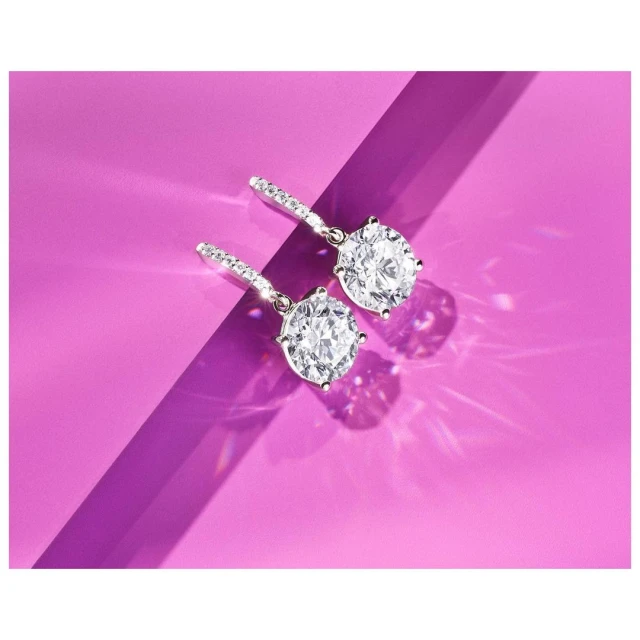Two round-cut diamond drop earrings are displayed on a purple background with visible shadows, highlighting their brilliant sparkle.