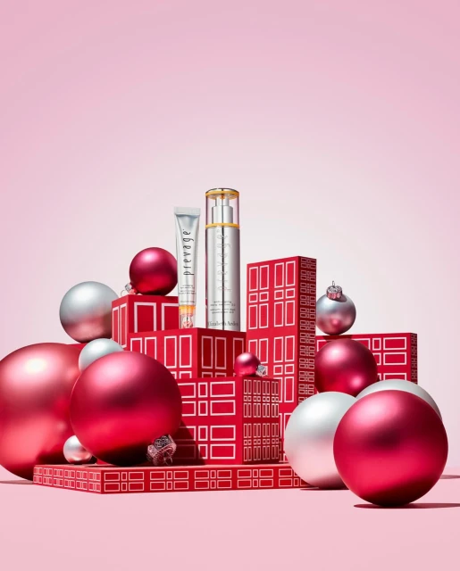 Two beauty product containers are displayed on a tiered platform with red gift boxes and silver and red ornaments against pink backdrop.