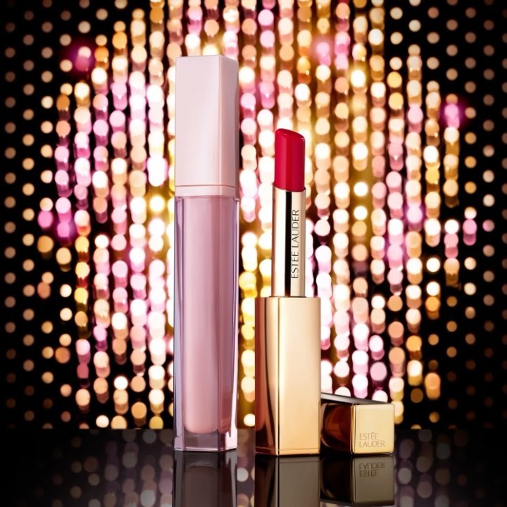 Estee Lauder lipstick and lip gloss in a beauty still life against a gold bokeh background on a black surface. Beauty product photography.