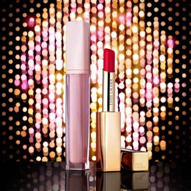 Estee Lauder lipstick and lip gloss in a beauty still life against a gold bokeh background on a black surface. Beauty product photography.