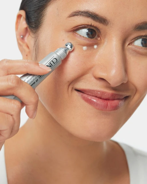 Close-up of woman applying Clinique eye cream with applicator under eye on clear skin against a white background in beauty shot.