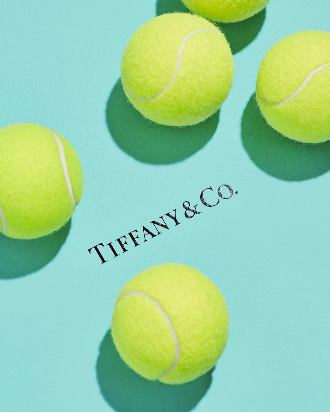 Elevated view of Tiffany & Co. branded box and scattered yellow tennis balls on a light blue background, creating a vibrant and sporty theme.