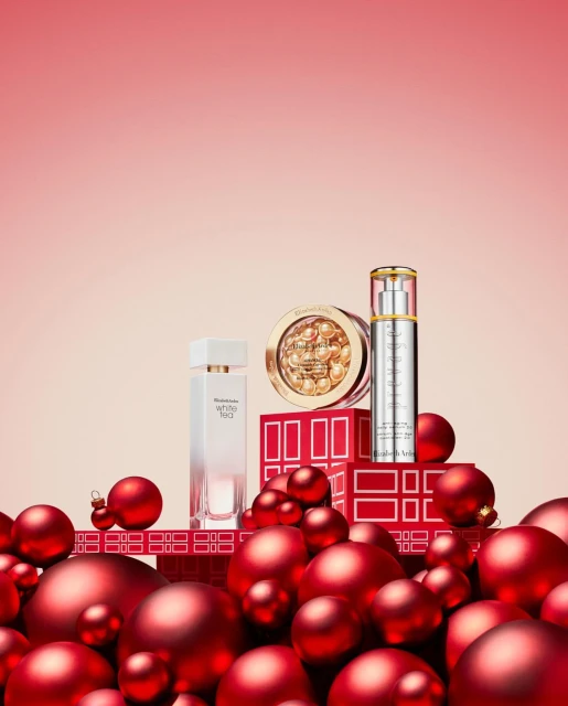 Still life shot of Elizabeth Arden beauty products arranged on red boxes, surrounded by red Christmas ornaments against a pink gradient backdrop.