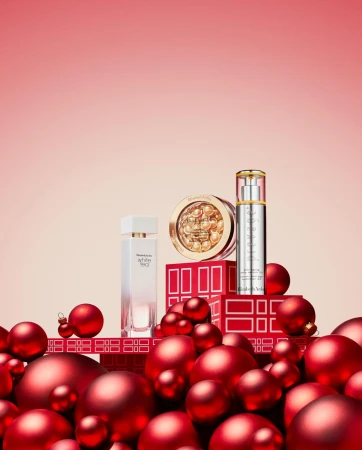 Still life shot of Elizabeth Arden beauty products arranged on red boxes, surrounded by red Christmas ornaments against a pink gradient backdrop.